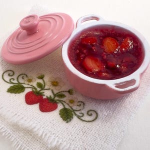 May include: A small pink ceramic pot with a lid, filled with strawberry jam. The pot sits on a white embroidered cloth with a strawberry design. The lid is off to the side.