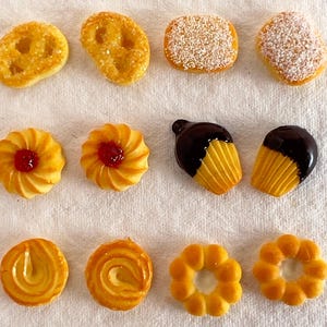 Puede incluir: Una variedad de galletas de diferentes formas y tamaños. Algunas están espolvoreadas con azúcar, otras tienen un diseño en espiral y algunas están cubiertas con una pizca de mermelada roja. Las galletas son de color marrón dorado y están dispuestas sobre una superficie blanca.