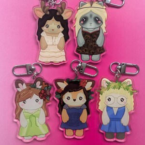K&ouml;nnte beinhalten: F&uuml;nf Acryl-Schl&uuml;sselanh&auml;nger mit illustrierten anthropomorphen Tierfiguren. Jede Figur tr&auml;gt ein anderes Outfit, darunter Kleider in Creme, Leopardenmuster, Hellgr&uuml;n, Dunkelblau und Blau, mit verschiedenen floralen und anderen Details.