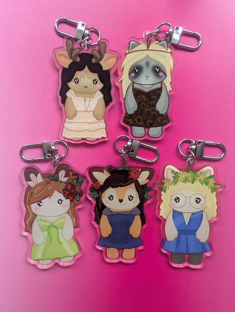 K&ouml;nnte beinhalten: F&uuml;nf Acryl-Schl&uuml;sselanh&auml;nger mit illustrierten anthropomorphen Tierfiguren. Jede Figur tr&auml;gt ein anderes Outfit, darunter Kleider in Creme, Leopardenmuster, Hellgr&uuml;n, Dunkelblau und Blau, mit verschiedenen floralen und anderen Details.