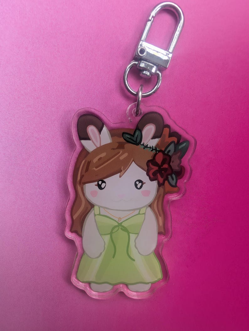K&ouml;nnte beinhalten: Ein Schl&uuml;sselanh&auml;nger aus klarem Acryl mit einer Zeichentrickfigur mit Hasenohren, braunem Haar und einem gr&uuml;nen Kleid. Die Figur tr&auml;gt ein dunkelrotes Blumenaccessoire im Haar und eine kleine goldene Halskette. Oben ist ein silberner Karabinerhaken aus Metall befestigt.