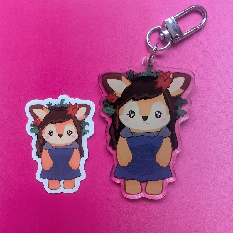 K&ouml;nnte beinhalten: Ein durchsichtiger Acryl-Schl&uuml;sselanh&auml;nger und ein passender Aufkleber mit einer Cartoon-Hirschfigur mit braunem Haar, einem blauen Kleid und einem Blumenkranz. Die Figur hat gro&szlig;e Augen, eine kleine herzf&ouml;rmige Nase und spitze Ohren.