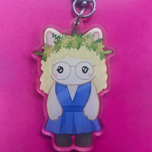 K&ouml;nnte beinhalten: Ein Acryl-Schl&uuml;sselanh&auml;nger mit einer Zeichentrickfigur mit blondem Haar, einem gr&uuml;nen Blumenkranz und einer runden Brille, die ein blaues Kleid tr&auml;gt. Die Figur hat wei&szlig;e Haut und ist an einem silbernen Karabinerhaken aus Metall befestigt.