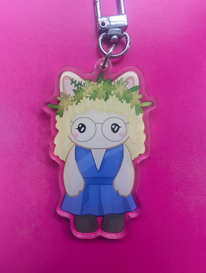 K&ouml;nnte beinhalten: Ein Acryl-Schl&uuml;sselanh&auml;nger mit einer Zeichentrickfigur mit blondem Haar, einem gr&uuml;nen Blumenkranz und einer runden Brille, die ein blaues Kleid tr&auml;gt. Die Figur hat wei&szlig;e Haut und ist an einem silbernen Karabinerhaken aus Metall befestigt.