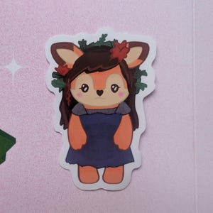 K&ouml;nnte beinhalten: Ein gestanzter Aufkleber mit einer Cartoon-Hirschfigur mit braunem Haar, die ein dunkelblaues Kleid und einen Kranz aus gr&uuml;nen Bl&auml;ttern und roten Blumen tr&auml;gt. Der Hirsch hat gro&szlig;e Augen, eine herzf&ouml;rmige Nase und rosa Wangen auf hellem Grund.