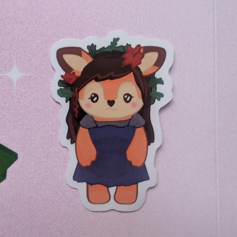 K&ouml;nnte beinhalten: Ein gestanzter Aufkleber mit einer Cartoon-Hirschfigur mit braunem Haar, die ein dunkelblaues Kleid und einen Kranz aus gr&uuml;nen Bl&auml;ttern und roten Blumen tr&auml;gt. Der Hirsch hat gro&szlig;e Augen, eine herzf&ouml;rmige Nase und rosa Wangen auf hellem Grund.