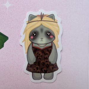K&ouml;nnte beinhalten: Ein gestanzter Aufkleber mit einem Cartoon-Waschb&auml;ren mit langem blondem Haar und einem d&uuml;nnen Stirnband. Der Waschb&auml;r hat gro&szlig;e schwarze Augen, rosa Wangen, einen neutralen Gesichtsausdruck und tr&auml;gt ein braunes Kleid mit Leopardenmuster.