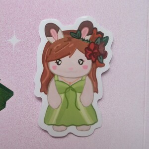 K&ouml;nnte beinhalten: Ein gestanzter Aufkleber mit einem anthropomorphen Hasencharakter mit braunem Haar, der ein hellgr&uuml;nes Kleid und einen Blumenkranz mit roten und dunkelgr&uuml;nen Bl&uuml;ten tr&auml;gt. Die Figur hat gro&szlig;e Augen, rosa Wangen und eine d&uuml;nne goldfarbene Halskette.