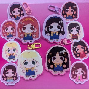 Può includere: Una collezione di portachiavi e adesivi di personaggi dei cartoni animati. I portachiavi hanno chiusure rosa, gialle e viola. Gli adesivi e i portachiavi raffigurano una ragazza dei cartoni animati con diversi colori di capelli, che indossa un vestito blu con un numero 2 giallo.