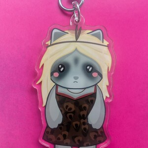 K&ouml;nnte beinhalten: Ein Acryl-Schl&uuml;sselanh&auml;nger mit einer Cartoon-Figur mit grauem Fell, gro&szlig;en dunklen Augen und langem blonden Haar. Die Figur tr&auml;gt ein braunes Kleid mit dunklem Leopardenmuster und ein schmales Stirnband. Der Anh&auml;nger hat einen silbernen Karabiner.