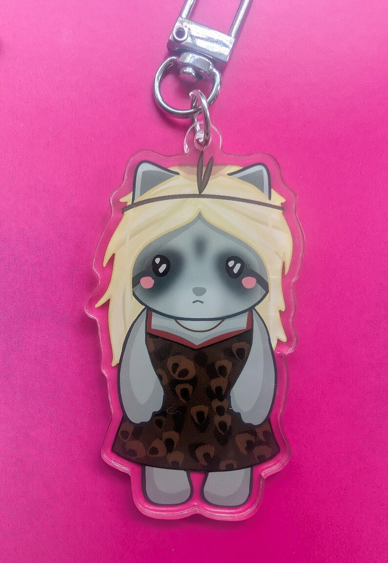 K&ouml;nnte beinhalten: Ein Acryl-Schl&uuml;sselanh&auml;nger mit einer Cartoon-Figur mit grauem Fell, gro&szlig;en dunklen Augen und langem blonden Haar. Die Figur tr&auml;gt ein braunes Kleid mit dunklem Leopardenmuster und ein schmales Stirnband. Der Anh&auml;nger hat einen silbernen Karabiner.