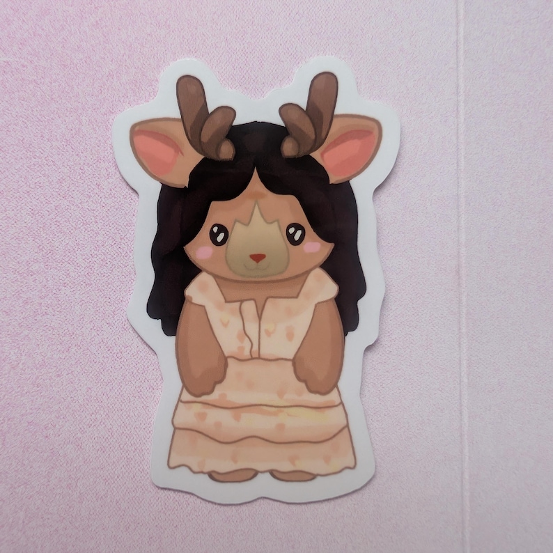 K&ouml;nnte beinhalten: Ein gestanzter Aufkleber mit einer Cartoon-Hirschfigur mit dunkelbraunem Haar und Geweih. Der Hirsch hat ein hellbraunes Gesicht mit rosa Wangen und tr&auml;gt ein blassgelbes, gestuftes Kleid mit einem dezenten Muster. Das Design hat einen wei&szlig;en Rand.