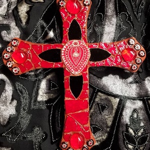 May include: A red and black mosaic cross with a white design in the center. The cross is decorated with red glass gems.