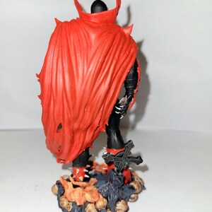 May include: A red and black action figure of a superhero with a cape and a sword. The figure is standing on a base with a skull design.