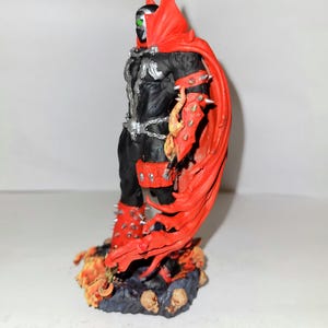 May include: A black and red action figure of Spawn, a comic book character, standing on a base with flames and skulls. The figure has a red cape and a green eye.
