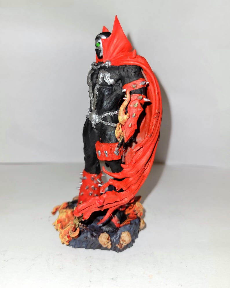 May include: A black and red action figure of Spawn, a comic book character, standing on a base with flames and skulls. The figure has a red cape and a green eye.