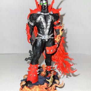 Puede incluir: Una figura de acción negra y roja de Spawn, un personaje de cómic, de pie sobre una base con llamas y calaveras. La figura tiene ojos verdes y una cadena plateada alrededor de la cintura.