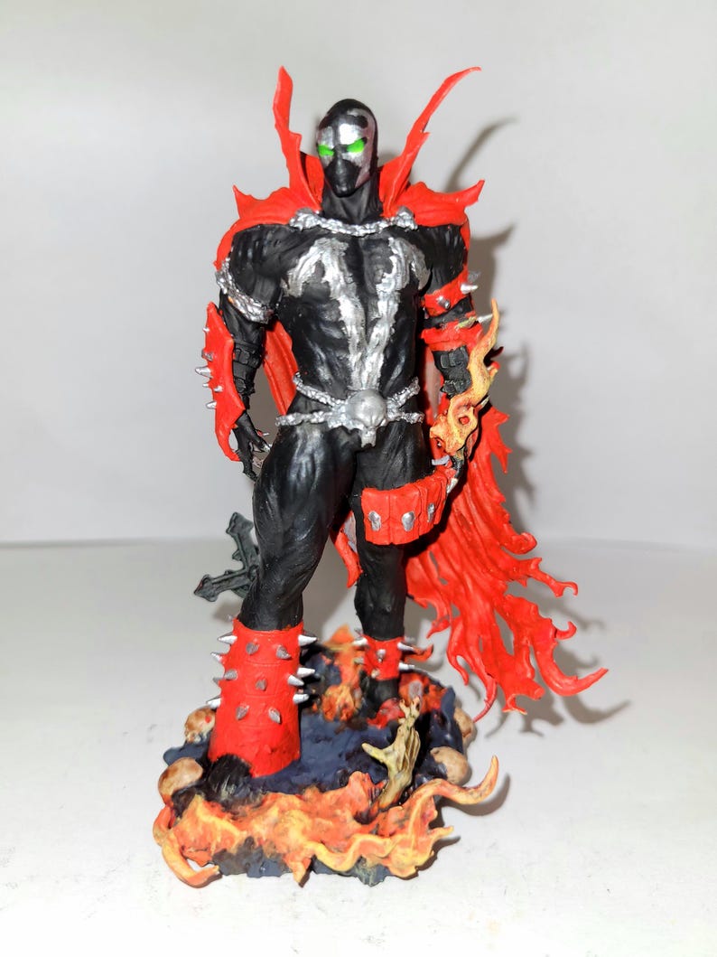 May include: A black and red action figure of Spawn, a comic book character, standing on a base with flames and skulls. The figure has green eyes and a silver chain around its waist.