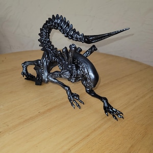 Puede incluir: Escultura detallada de una criatura alienígena con aspecto metálico, con una larga cola segmentada y múltiples extremidades. La escultura es de color gris oscuro y tiene una superficie texturizada, garras afiladas y una pose amenazante.