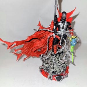 Puede incluir: Una figura de acción detallada de Spawn, un personaje de cómic, con una capa roja y un bastón plateado. La figura está de pie sobre una base con un diseño de calavera.