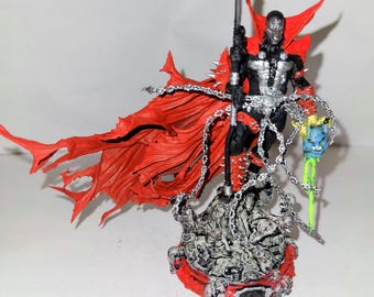 May include: A detailed action figure of Spawn, a comic book character, with a red cape and a silver staff. The figure is standing on a base with a skull design.