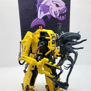 May include: A yellow and black mech suit toy with a figure inside, facing a black Alien figure. A purple fish graphic is in the background. The mech has a light on top and black stripes.