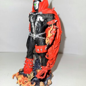May include: A black and red action figure of Spawn, a comic book character, standing on a base with flames and skulls. The figure has a cape, spiked armour, and a skull-shaped belt.