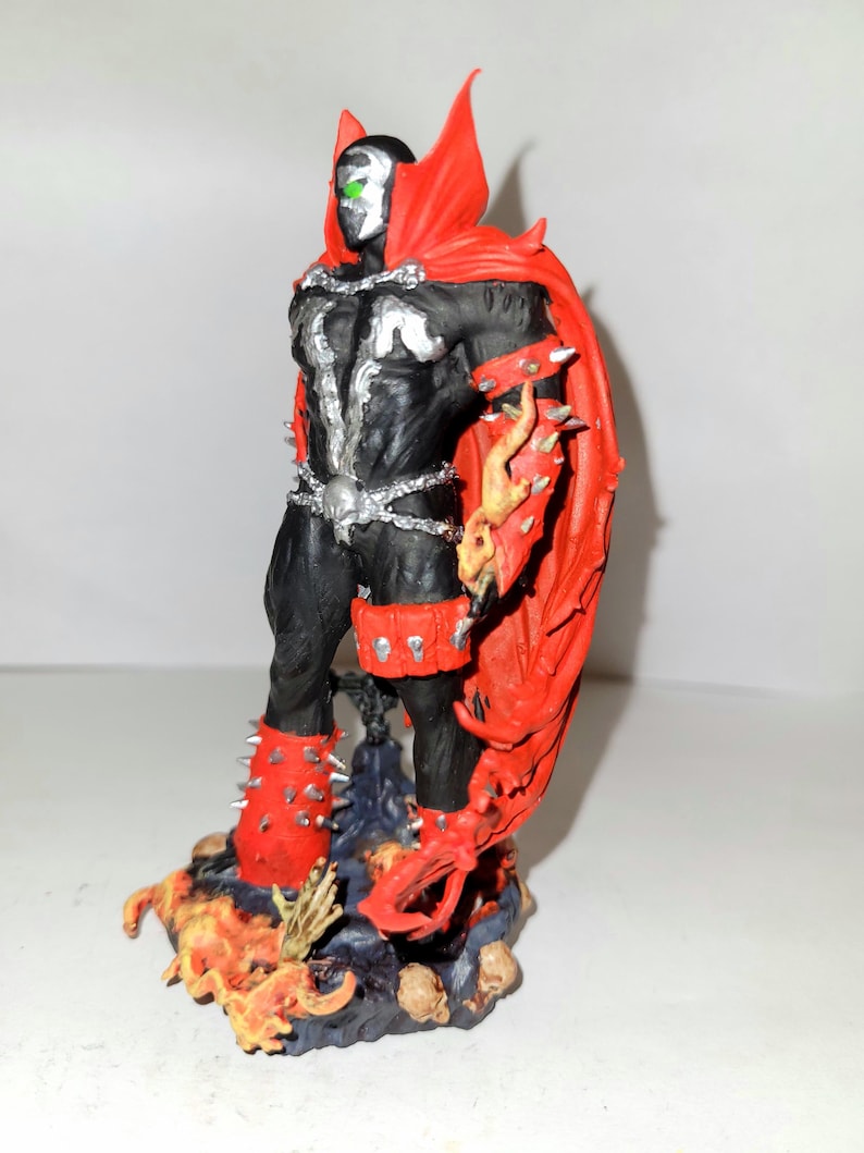 May include: A black and red action figure of Spawn, a comic book character, standing on a base with flames and skulls. The figure has a cape, spiked armour, and a skull-shaped belt.
