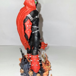 May include: A red and black action figure of Spawn, a superhero character, standing on a base with flames and skulls. The figure has a red cape and spiked boots.