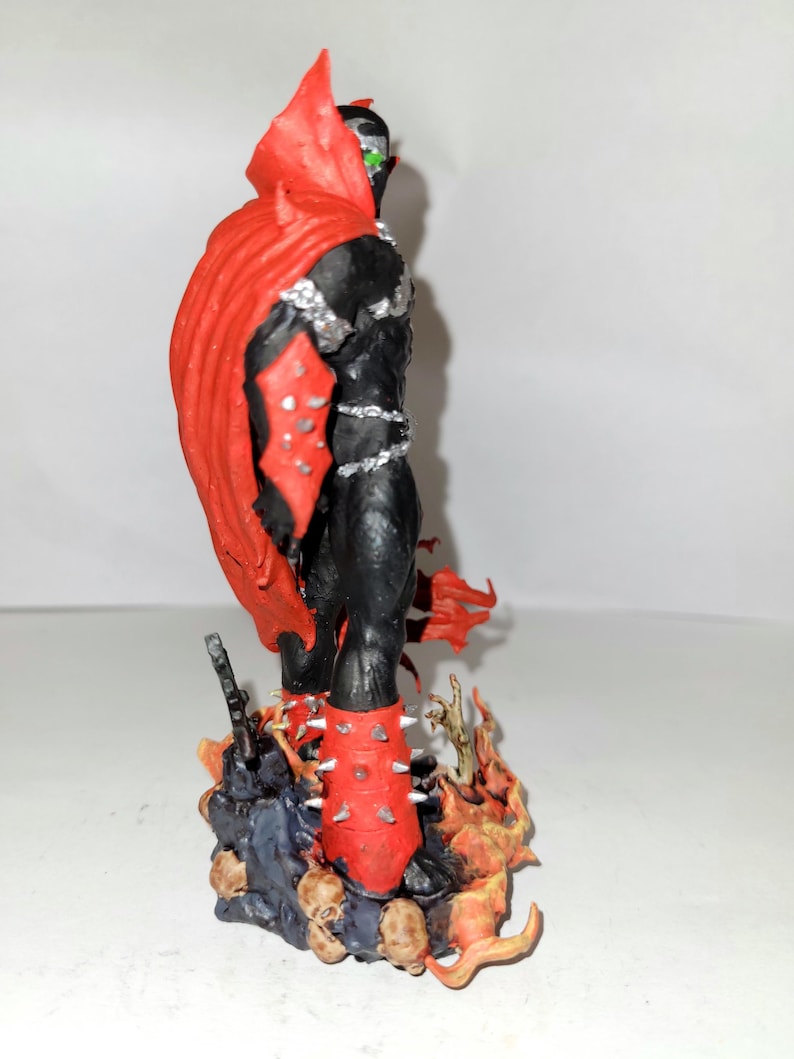 May include: A red and black action figure of Spawn, a superhero character, standing on a base with flames and skulls. The figure has a red cape and spiked boots.