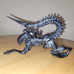 Pu&ograve; includere: Una statuetta di una creatura aliena color argento con una lunga coda segmentata e una testa appuntita. La creatura &egrave; in posizione accovacciata con la testa rivolta verso il basso e la coda arrotolata dietro di lei.