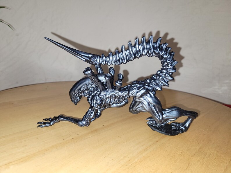 Pu&ograve; includere: Una statuetta di una creatura aliena color argento con una lunga coda segmentata e una testa appuntita. La creatura &egrave; in posizione accovacciata con la testa rivolta verso il basso e la coda arrotolata dietro di lei.