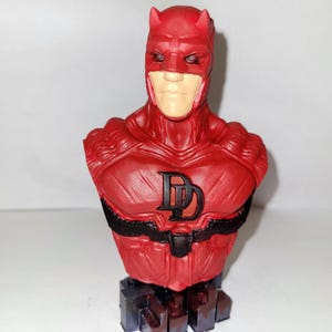May include: A red and black Daredevil bust figurine. The bust is made of plastic and features a detailed sculpt of Daredevil's head and torso. The bust is mounted on a black base.