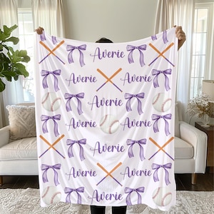 May include: White baseball-themed blanket with a repeating pattern of baseballs, crossed bats, and purple bows. The name "Averie" is written in purple script throughout the design. The blanket is held up by a person.