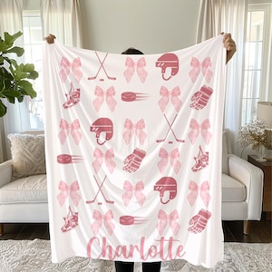May include: White blanket with a pink hockey theme, featuring hockey helmets, sticks, pucks, skates, and bows. The name "Charlotte" is printed in pink script at the bottom. The blanket is held up in front of a white couch and window.