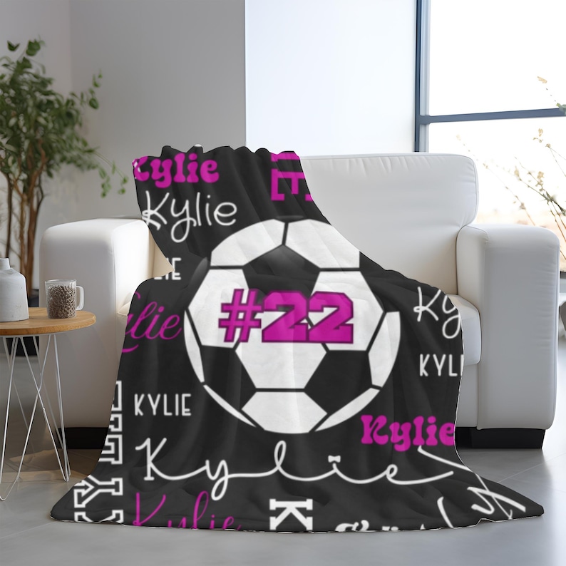 May include: Black fleece blanket with a soccer ball graphic and the name "Kylie" in white and pink lettering. The soccer ball has the number "22" in pink. The blanket is draped over a white couch.