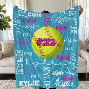 May include: A blue and white softball themed fleece blanket with the name "Kylie" repeated throughout the design. A yellow softball with red stitching and the number "22" in pink is centered on the blanket.