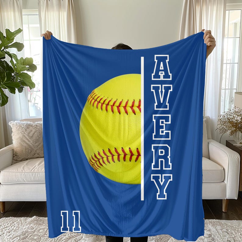 May include: A blue blanket featuring a large yellow softball with red stitching. The name "AVERY" is printed vertically in white, along with the number "11" at the bottom. The blanket is held up in front of a person.