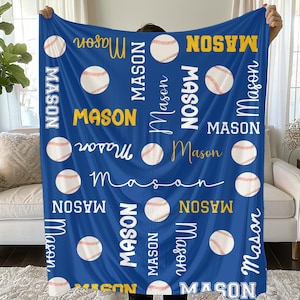 Op de afbeelding: Een blauwe deken met honkballen en de naam "Mason" in verschillende lettertypen en kleuren. Het ontwerp bevat witte honkballen met rode stiksels, en de naam is in wit en geel. De deken wordt door een persoon omhoog gehouden.