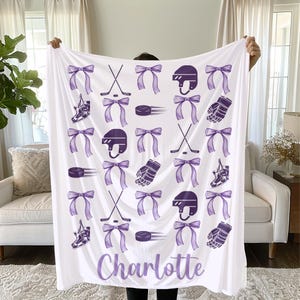 May include: White hockey-themed blanket featuring purple bows, hockey sticks, helmets, pucks, skates, and gloves. The name "Charlotte" is written in purple script at the bottom. The blanket is held up by a person.