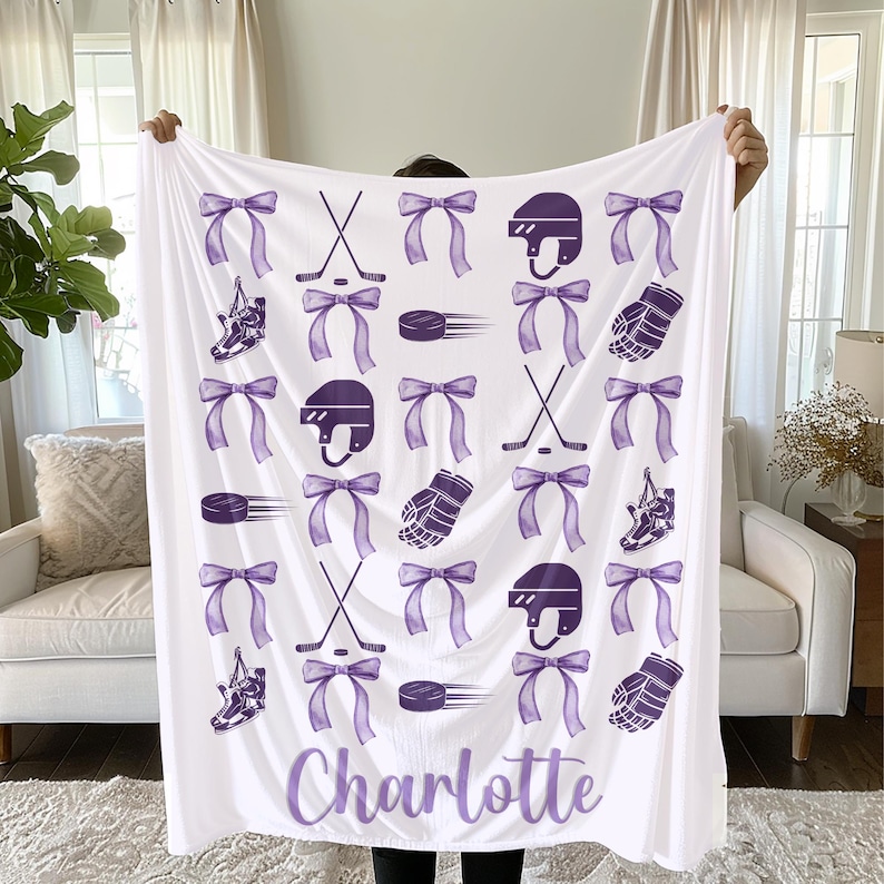 May include: White hockey-themed blanket featuring purple bows, hockey sticks, helmets, pucks, skates, and gloves. The name "Charlotte" is written in purple script at the bottom. The blanket is held up by a person.