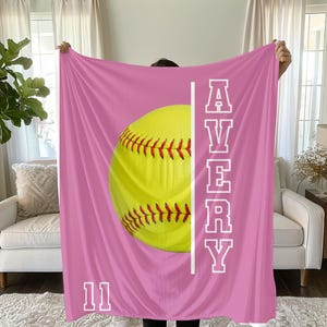 May include: A pink softball blanket featuring a large yellow softball with red stitching. The name "AVERY" is printed vertically in white, along with the number "11" at the bottom. The blanket is held up by a person.