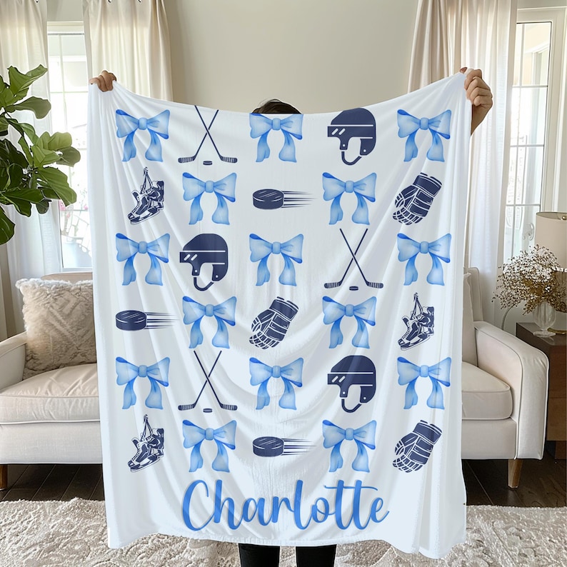 May include: White hockey-themed blanket with blue bows, hockey sticks, helmets, gloves, and skates. The name "Charlotte" is printed in blue script at the bottom. The blanket is held up by a person.