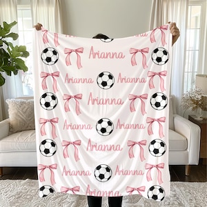 May include: White blanket with a repeating pattern of soccer balls, pink bows, and the name "Arianna" in pink script. The blanket is held up, showcasing the design.