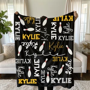 以下が含まれることがあります： 白と金の文字で「Kylie」という名前と白いボーリングピンが描かれた黒いブランケット。パーソナライズされたアイテムで、家の装飾に適しています。ブランケットのデザインには様々なフォントが含まれています。