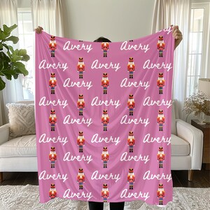 May include: A pink blanket featuring a repeating pattern of toy nutcrackers in red, gold, and black uniforms. The name "Avery" is written in white script across the blanket. The blanket is held up by a person.