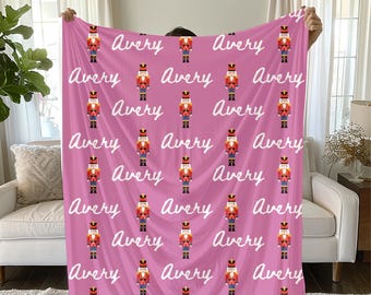 Puede incluir: Una manta rosa con un patrón repetido de cascanueces de juguete con uniformes rojos, dorados y negros. El nombre "Avery" está escrito en blanco en la manta. La manta está sostenida por una persona.
