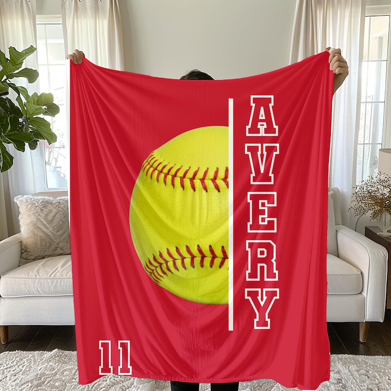 May include: A red blanket featuring a large yellow softball with red stitching. The name "AVERY" is printed vertically in white, along with the number "11". The blanket is a sports-themed item.