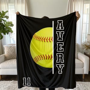 May include: Black blanket featuring a large yellow softball with red stitching. The name "AVERY" is printed vertically in white block letters, with the number "11" at the bottom. The blanket is held up by a person.