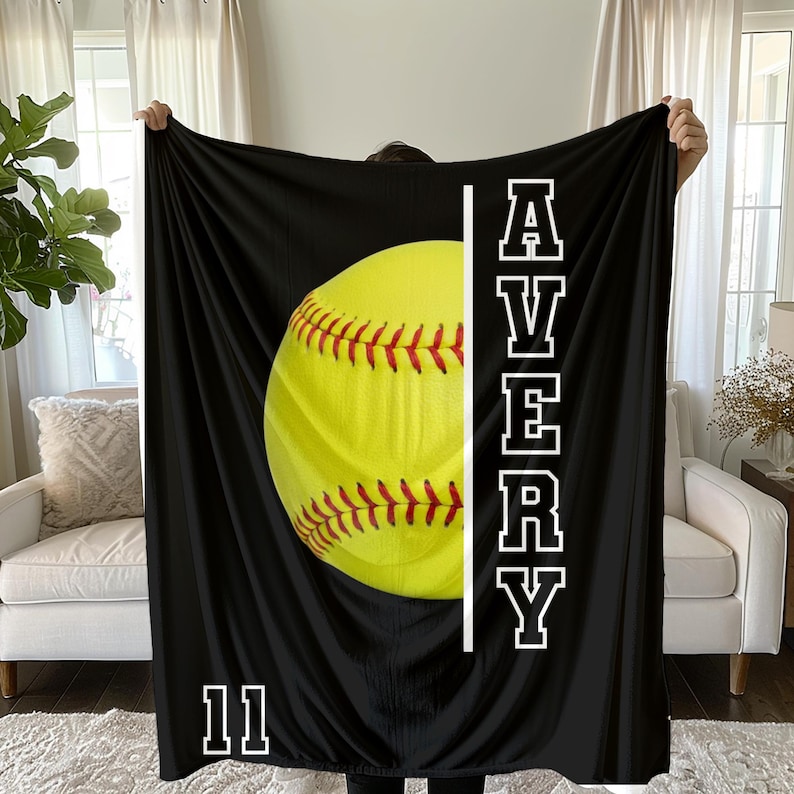 May include: Black blanket featuring a large yellow softball with red stitching. The name "AVERY" is printed vertically in white block letters, with the number "11" at the bottom. The blanket is held up by a person.